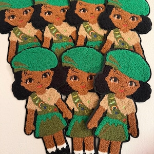 May include: Seven identical embroidered patches featuring a cartoon character with dark skin, black hair, and a green beret. The character wears a tan shirt, a green skirt, and a green sash decorated with a rainbow, a star, and circular badges.
