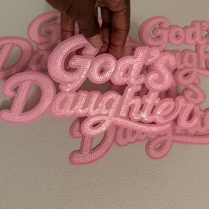 May include: A collection of light pink sequin patches featuring the text God's Daughter in a cursive script font. Each patch is crafted with individual sequins that create a textured, shimmering surface, designed for application onto fabric items.
