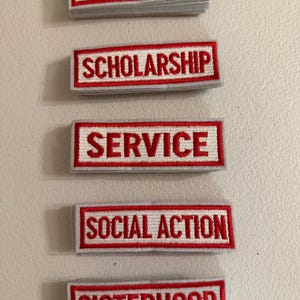May include: Five rectangular embroidered patches with white backgrounds and red borders. Each patch features red embroidered text reading FORTITUDE, SCHOLARSHIP, SERVICE, SOCIAL ACTION, and SISTERHOOD, stacked vertically in a column.