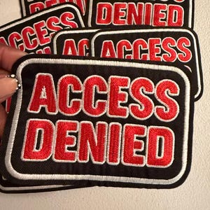 May include: A rectangular embroidered patch with a black background and a white border. The text ACCESS DENIED is stitched in bold red capital letters across the center. The patch features a thick, raised thread texture throughout the design.