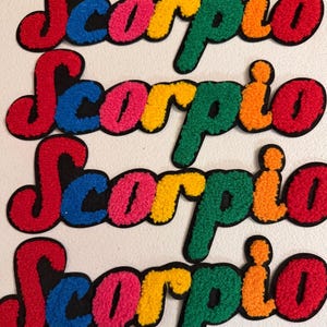 May include: Four identical embroidered patches featuring the word Scorpio in a cursive font. Each letter is a different color, including red, blue, pink, yellow, green, and orange, with a black outline and a textured, fuzzy surface finish.
