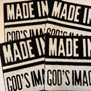 May include: Four black and white rectangular towels. Each towel has the words "MADE IN" in bold white letters at the top, and "GOD'S IMAGE" in white letters at the bottom. The towels are stacked on top of each other.