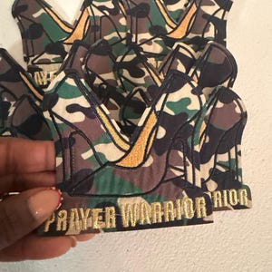 May include: Camouflage-patterned crown patches with gold embroidery. The patches feature a high-heeled shoe design and the words "PRAYER WARRIOR" embroidered in gold. The patches are in a pile, with one held up.