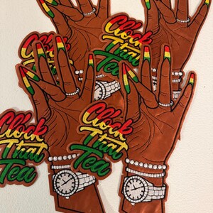 May include: Multiple embroidered patches featuring a brown-toned hand with long nails painted in red, yellow, and green. The hand wears a silver ring, a beaded bracelet, and a silver wristwatch. The text Clock That Tea is embroidered in red, yellow, and green.