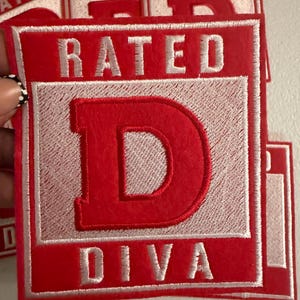 May include: A rectangular embroidered patch featuring a red border and a white center square. The word RATED is stitched in white at the top, a large red letter D is centered in the middle, and the word DIVA is stitched in white at the bottom.