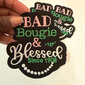 May include: Two black embroidered patches with scalloped edges. The text reads BAD in pink, Bougie in green, & in pink, and Blessed in white. Below, the text reads Since 1908 in green, accompanied by a white embroidered pearl necklace design.