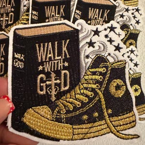 May include: An embroidered patch featuring a black book with the gold text WALK WITH GOD and a cross symbol, alongside a black high-top sneaker with gold laces and trim, accented by swirling gray smoke and small black stars on a white background.