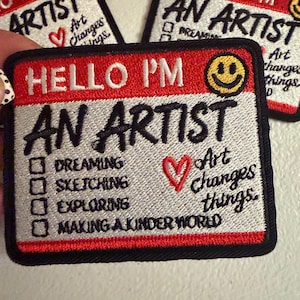 May include: Rectangular embroidered patches featuring a red and white name tag design. The text reads HELLO I'M AN ARTIST with a yellow smiley face. Below are checkboxes for DREAMING, SKETCHING, EXPLORING, MAKING A KINDER WORLD and the text Art changes things.