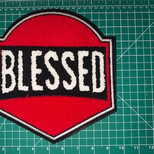 May include: A red and black patch with the word "BLESSED" in white, bold letters. The patch has a unique shape with a black border and a white outline. The background is a green cutting mat.