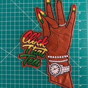 K&ouml;nnte beinhalten: Ein gestickter Aufn&auml;her, der eine braun get&ouml;nte Hand mit langen, in Rot, Gelb und Gr&uuml;n lackierten Fingern&auml;geln zeigt. Die Hand tr&auml;gt einen silbernen Ring, Armb&auml;nder und eine Uhr. Der Text "Clock That Tea" ist in rotem, gelbem und gr&uuml;nem Garn gestickt.