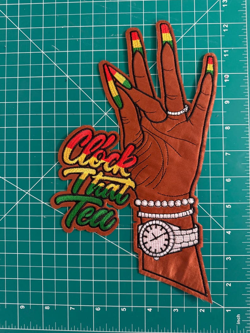 K&ouml;nnte beinhalten: Ein gestickter Aufn&auml;her, der eine braun get&ouml;nte Hand mit langen, in Rot, Gelb und Gr&uuml;n lackierten Fingern&auml;geln zeigt. Die Hand tr&auml;gt einen silbernen Ring, Armb&auml;nder und eine Uhr. Der Text "Clock That Tea" ist in rotem, gelbem und gr&uuml;nem Garn gestickt.