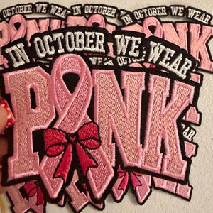 May include: An embroidered patch featuring the text IN OCTOBER WE WEAR PINK in white letters on a black background. The word PINK is stylized with a light pink awareness ribbon forming the letter I and a dark pink bow accenting the ribbon.