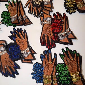 May include: Embroidered patches featuring brown hands adorned with rings, bracelets, and watches. The nails are painted in various colors, including blue, red, and green. Each patch has the text "Clock That Tea" in a matching color.