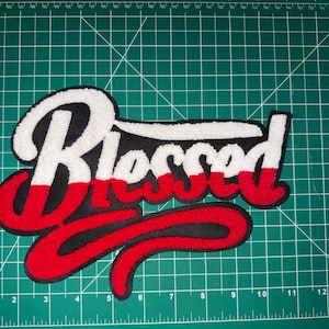 Puede incluir: Un parche bordado grande con la palabra "Blessed" en una escritura estilizada. La rotulaci&oacute;n es principalmente blanca, roja y negra, con una apariencia texturizada y gruesa. El parche est&aacute; probablemente dise&ntilde;ado para su aplicaci&oacute;n en ropa o accesorios.