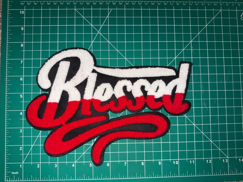Puede incluir: Un parche bordado grande con la palabra "Blessed" en una escritura estilizada. La rotulaci&oacute;n es principalmente blanca, roja y negra, con una apariencia texturizada y gruesa. El parche est&aacute; probablemente dise&ntilde;ado para su aplicaci&oacute;n en ropa o accesorios.