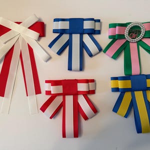 May include: A collection of six decorative ribbon bows in various color combinations. The bows feature red, white, blue, green, pink, and yellow ribbons. One bow has a silver embellishment with the text "Pretty Girl".