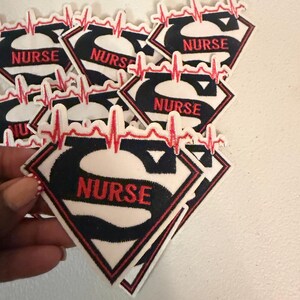 May include: A collection of embroidered patches shaped like a diamond shield with a black letter S, the word NURSE in red thread, and a red EKG heartbeat line across the top. Each patch features a white background with a black and red border.