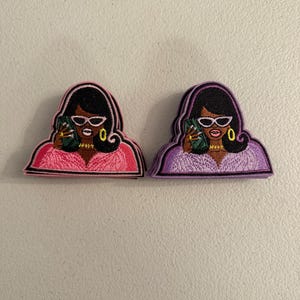 May include: Two embroidered patches featuring a stylized figure with dark hair, white cat-eye sunglasses, gold hoop earrings, and a gold necklace. One patch shows the figure in a bright pink top holding a green clutch, while the other shows a lavender top.