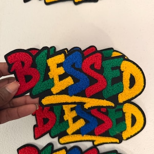 May include: A stack of embroidered patches featuring the word BLESSED in a graffiti-style font. Each letter is a different color, including red, green, blue, and yellow, with a black outline. The texture of the embroidery is raised and visible.