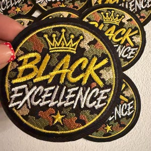 May include: A circular embroidered patch featuring a camouflage background in shades of green, brown, and tan. The patch displays a yellow embroidered crown above the text "BLACK EXCELLENCE" in yellow and white thread, with a small yellow star.