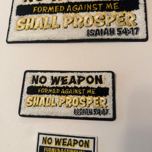May include: Three embroidered patches with the text "NO WEAPON FORMED AGAINST ME SHALL PROSPER ISAIAH 54:17". The patches are rectangular, with black and yellow lettering on a white background. The largest patch has a textured appearance.