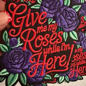 May include: A black embroidered patch featuring the red stitched text Give me my Roses while I'm Here. The design is decorated with purple roses and light green leaves. The embroidery is dense and textured, creating a clear contrast against the black fabric background of the patch.