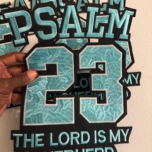 May include: A black and light blue embroidered patch featuring the word PSALM at the top, the number 23 in the center, and the text THE LORD IS MY SHEPHERD at the bottom. The design uses a textured light blue thread on a black fabric background.