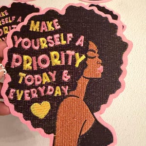 May include: An embroidered patch featuring a profile silhouette of a person with dark skin and voluminous black hair. The text reads MAKE YOURSELF A PRIORITY TODAY & EVERYDAY in pink and yellow thread, accompanied by a small yellow heart.