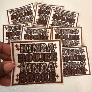May include: A collection of square patches with the text "KINDA BOUJEE KINDA BROKE" in brown leopard print lettering. The patches have a white background and a brown stitched border. Some patches are held by a hand.