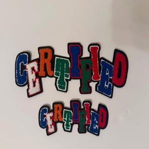 May include: Two sets of colorful, embroidered patches spelling "CERTIFIED". The patches feature a variety of colors including blue, orange, green, and red, with white and black accents. The patches are designed with a varsity letter style.