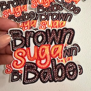 May include: A collection of embroidered patches featuring the text Brown Suga Babe in a stacked layout. The words Brown and Babe are stitched in dark brown thread with white accents, while the word Suga is in bright orange and red thread.