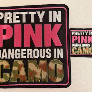 May include: Two square embroidered patches with black borders and pink trim. The text reads PRETTY IN PINK DANGEROUS IN CAMO. The word PINK is in bright pink thread, DANGEROUS IN is in white, and CAMO is in a green, brown, and tan camouflage pattern.