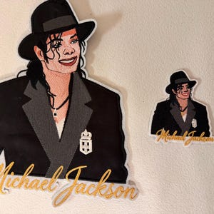 May include: Two embroidered patches featuring a portrait of Michael Jackson wearing a black fedora and a black blazer with grey lapels. The larger patch includes a white crest emblem on the lapel and the name Michael Jackson in gold script.