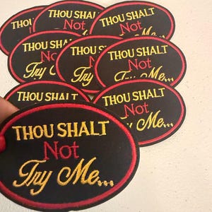 May include: A collection of black oval patches with red borders. Each patch features the embroidered phrase "THOU SHALT Not Try Me..." in gold and red thread, creating a bold statement.