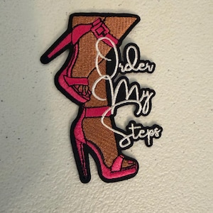 May include: An embroidered patch featuring two legs wearing bright pink high-heeled sandals with ankle straps. The design includes the white cursive text Order My Steps layered over the legs, all outlined in black thread on a light background.