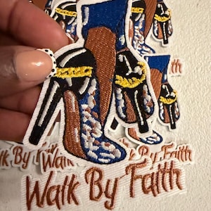 May include: An embroidered patch featuring a high-heeled shoe with blue, black, and gold accents, worn on a brown-toned leg. The patch includes the text "Walk By Faith" in brown script lettering, with a white border surrounding the entire design.
