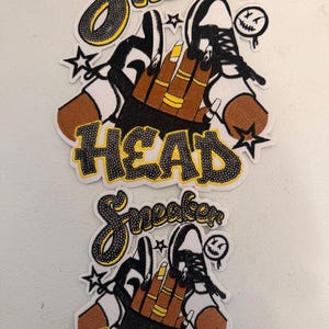 May include: Two embroidered patches with the words "Sneaker Head" in black and yellow script. The patches feature a hand with gold rings and yellow nails, a black and white sneaker, and a small, smiling face.