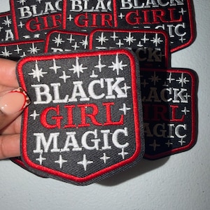 May include: Black Girl Magic patches with a shield shape. The patches have a black background with white text and a red border. The words "BLACK GIRL MAGIC" are embroidered on the patch.