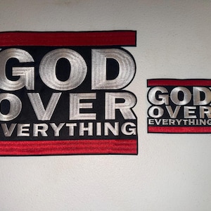 May include: Two embroidered patches with the text "GOD OVER EVERYTHING". The larger patch has silver lettering on a black background, framed by red borders. The smaller patch mirrors the design. Both are displayed against a white backdrop.