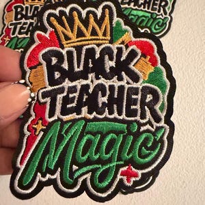 May include: An embroidered patch featuring the text Black Teacher Magic in black and green thread. A gold crown sits above the text, accented by red, yellow, and green embroidery. The design is outlined in white and black with a black border.