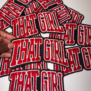 May include: A collection of red, black, and white embroidered patches. Each patch features the phrase "THAT GIRL" in a bold, collegiate-style font. The patches are piled together, with one being held by a hand.