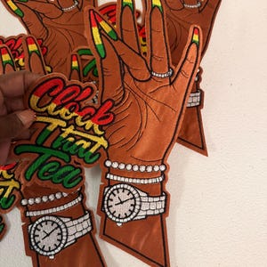 K&ouml;nnte beinhalten: Ein gestickter Aufn&auml;her mit einer braun get&ouml;nten Hand mit langen, rot, gelb und gr&uuml;n lackierten Fingern&auml;geln. Die Hand tr&auml;gt einen Ring, ein Perlenarmband und eine Armbanduhr. Der Text "Clock That Tea" ist in Rot, Gelb und Gr&uuml;n gestickt.