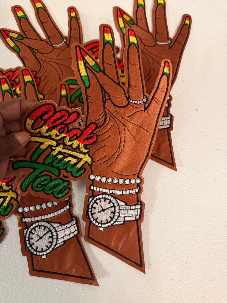 K&ouml;nnte beinhalten: Ein gestickter Aufn&auml;her mit einer braun get&ouml;nten Hand mit langen, rot, gelb und gr&uuml;n lackierten Fingern&auml;geln. Die Hand tr&auml;gt einen Ring, ein Perlenarmband und eine Armbanduhr. Der Text "Clock That Tea" ist in Rot, Gelb und Gr&uuml;n gestickt.