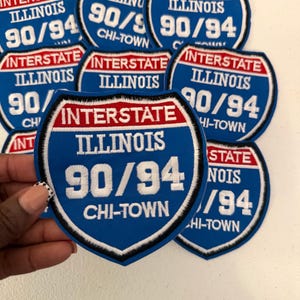 May include: A collection of shield-shaped embroidered patches featuring a blue background with a red top banner. The text reads INTERSTATE, ILLINOIS, 90/94, and CHI-TOWN in white lettering. The patches have a black and white border design.