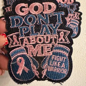 May include: An embroidered patch with a black background featuring the pink and blue text "GOD DON'T PLAY ABOUT ME". The design includes two boxing gloves, one with a pink awareness ribbon and the other with the text "FIGHT LIKE A WARRIOR".
