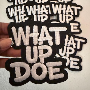 May include: A collection of black embroidered patches featuring the white text "WHAT UP DOE" in a bold, sans-serif font. Each patch has a thick black border and a raised, textured stitch finish, creating a clear contrast between the white lettering and the black fabric background.