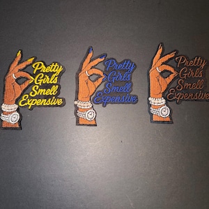 May include: Three embroidered patches with a hand gesture and the text "Pretty Girls Smell Expensive." Each patch features a different color scheme: yellow, blue, and orange. The hands have manicured nails and are adorned with jewelry.