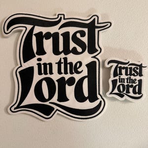 May include: Two die-cut stickers featuring the black text Trust in the Lord in a bold, stylized font. The larger sticker is on the left and the smaller version is on the right. Both stickers have a white border and a white background.