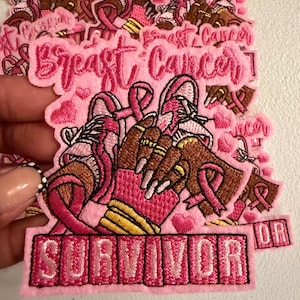 May include: An embroidered patch featuring a pair of pink boxing gloves, a pink ribbon, and the text Breast Cancer SURVIVOR. The design includes small pink hearts and a hand wearing a pink wrist wrap and gold rings, all rendered in shades of pink.