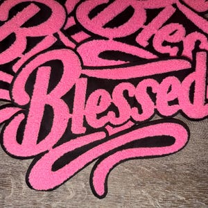 Puede incluir: Primer plano de un parche rosa y negro con la palabra "Blessed" en cursiva. El parche tiene una textura difusa y est&aacute; sobre un fondo de madera. La palabra "Blessed" es el foco principal.